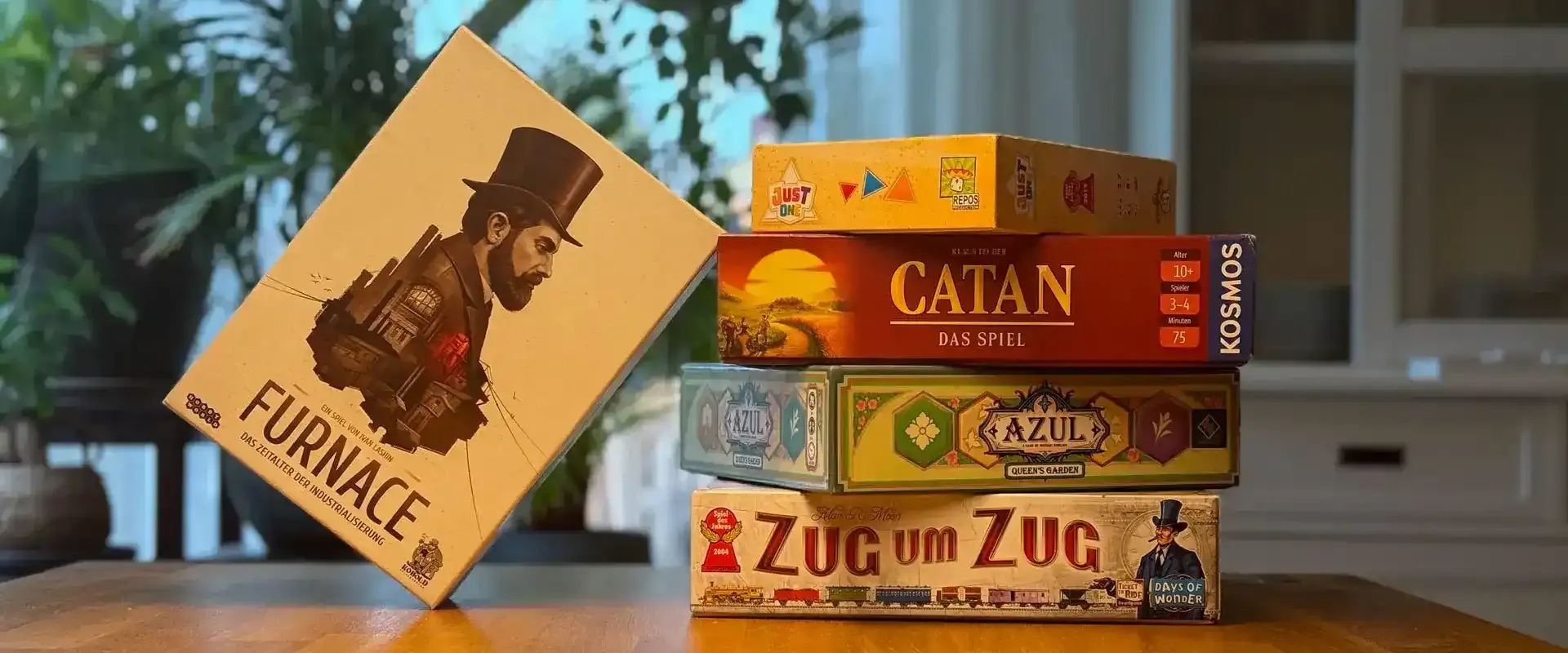 Auf dem Bild ist ein Holztisch. Im Hintergrund sind Pflanzen zu sehen. Auf dem Tisch liegen fünf Spiele: “Zug um Zug”, “Azul”, “Siedler von Catan”, “Just One” und “Furnace”. Vier davon sind aufeinandergestapelt, das fünfte lehnt von links am Stapel.